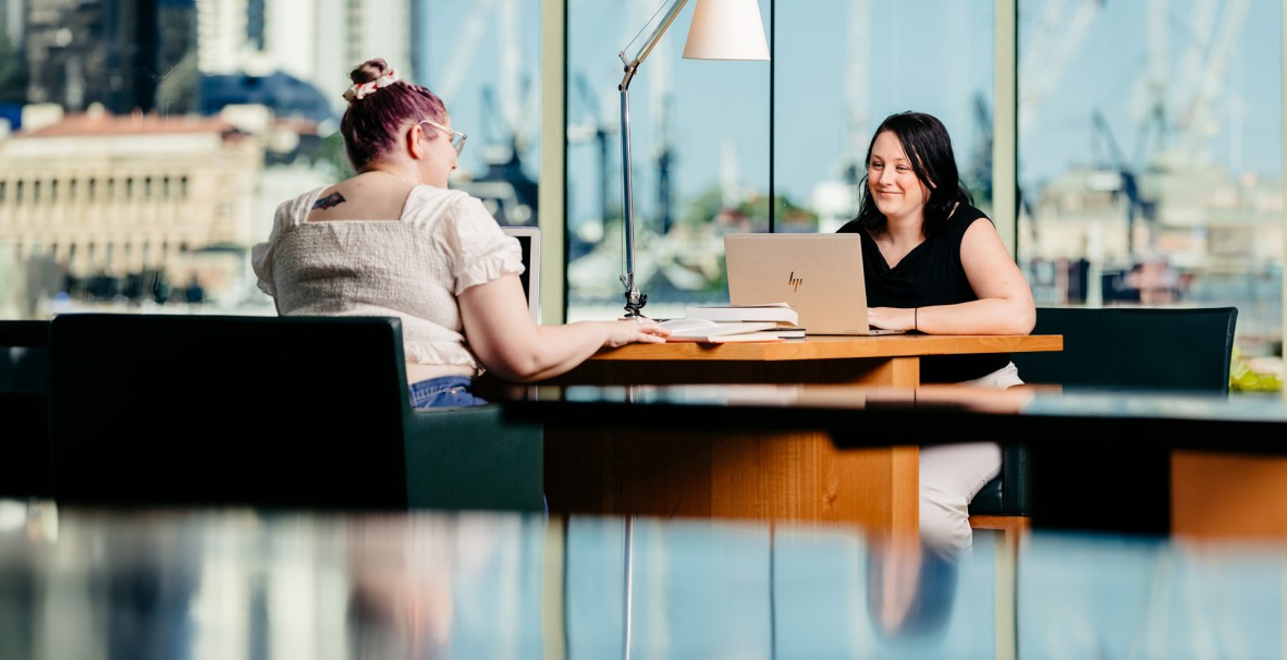 Two women sitting at a joint desk with a view of Brisbane city using laptops at State Library.