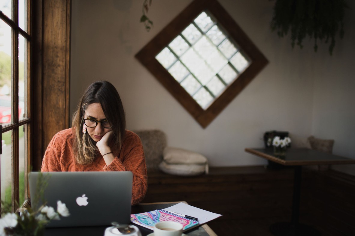 Kirli Saunders sits at a Mac laptop in a cafe. She wears a dark orange jumper and her hair is down. She is typing. 