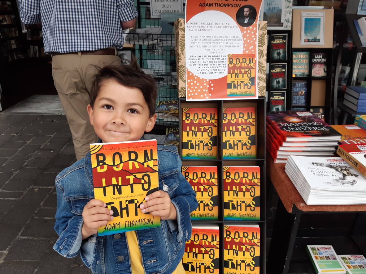 An adorable little boy holds up a copy of Born into this He stands at a bookshop in front of a big display of the book 