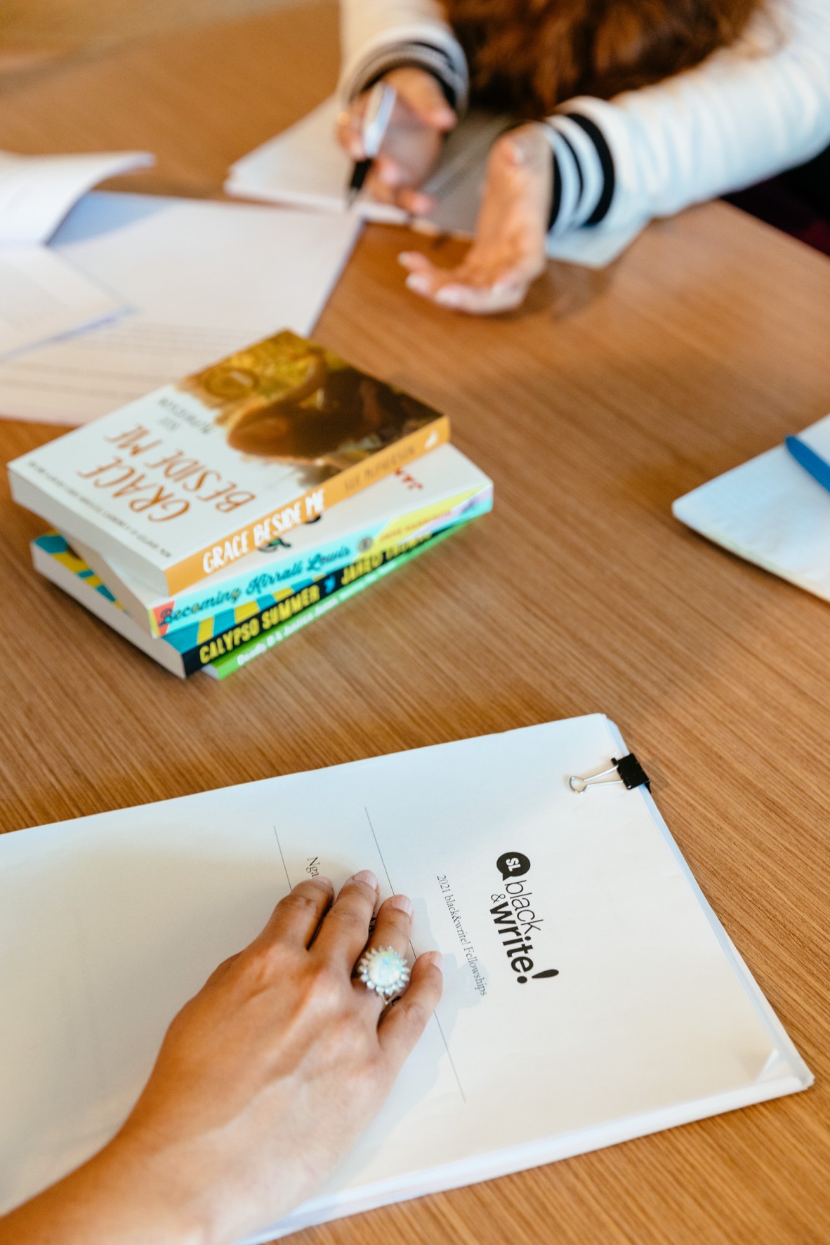 Mid shot of a table with black&write! books and manuscripts on it. People's hands are visible, they look like they are having a conversation about stories.
