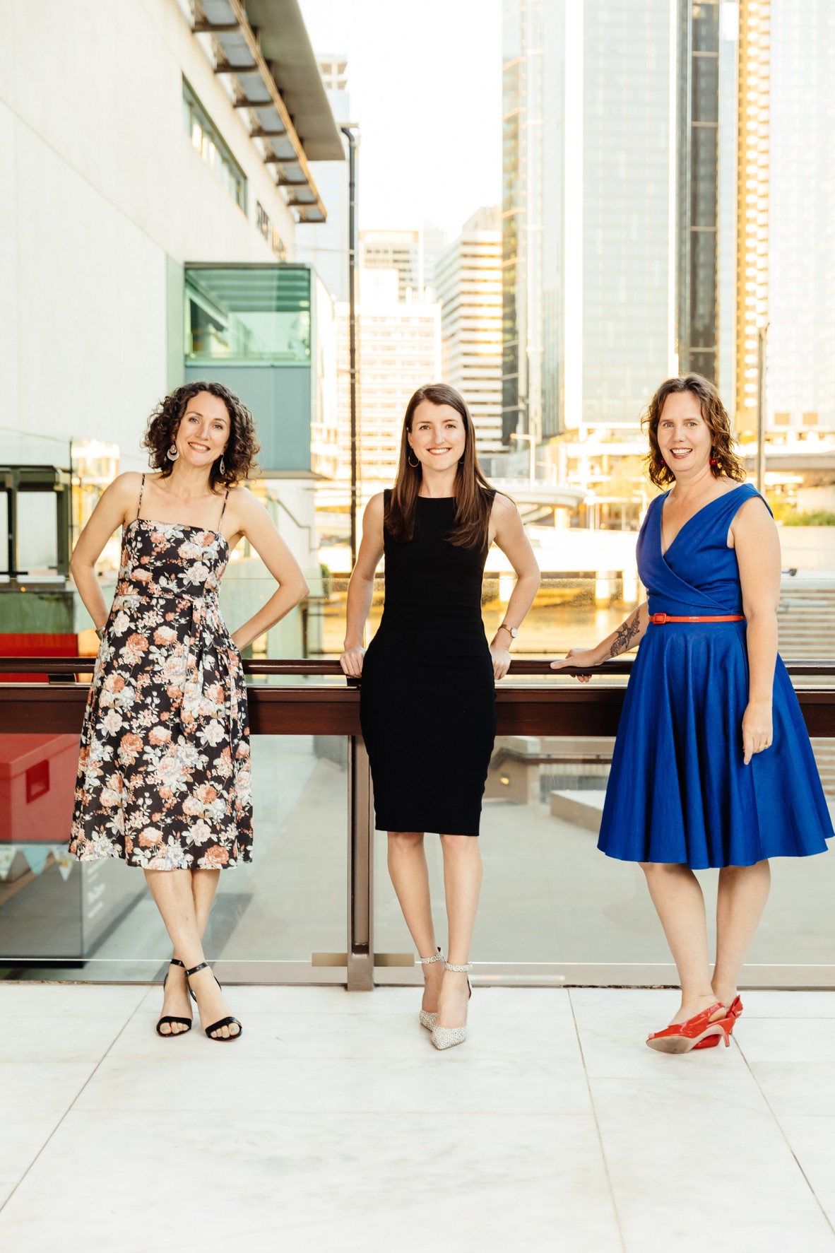 Kali Napier in a floral dress, Ella Jeffery in black and Tabitha Bird in blue stand smiling at the railing of the Queensland Terrace