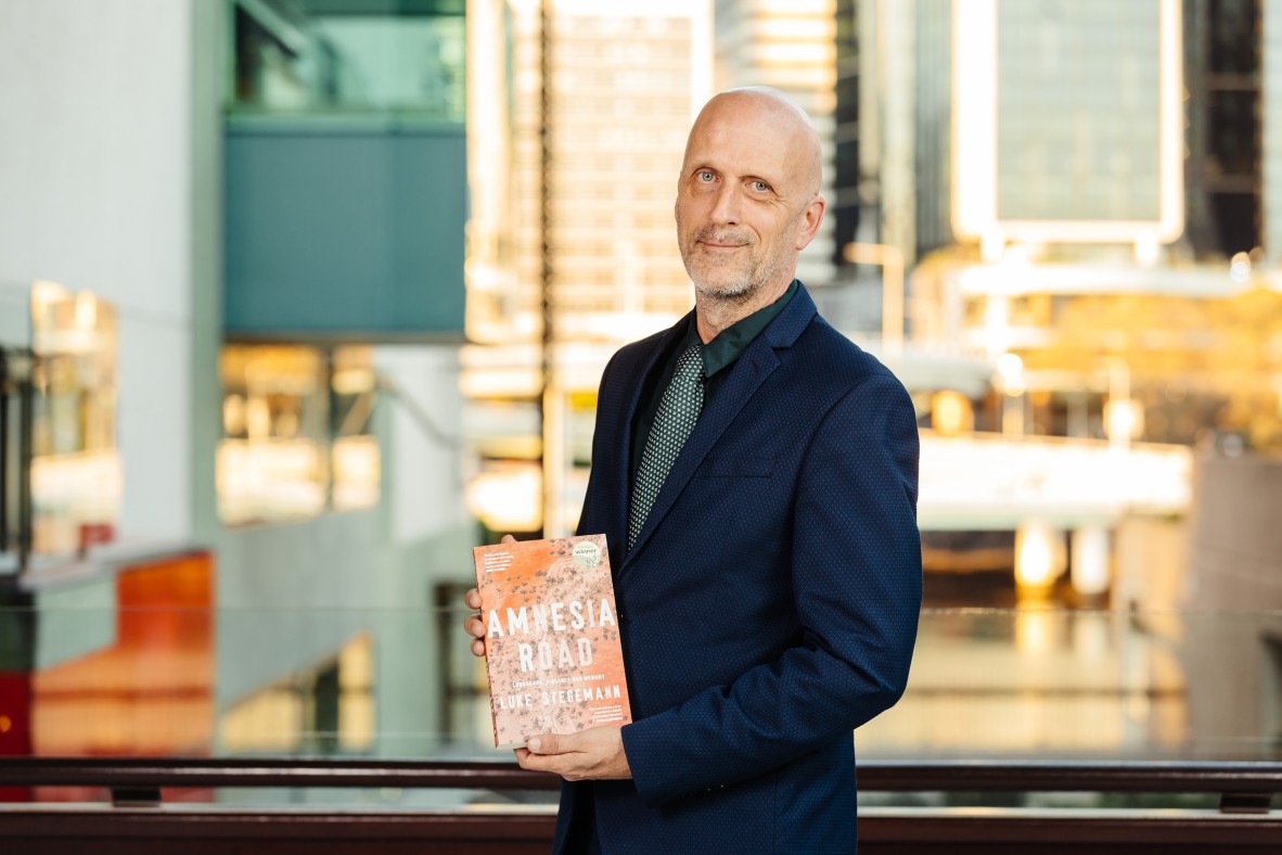 Luke Stegemann stands at sunset on the Queensland Terrace. He holds a copy of his book Amnesia Road, at the2021 Queensland Literary Awards