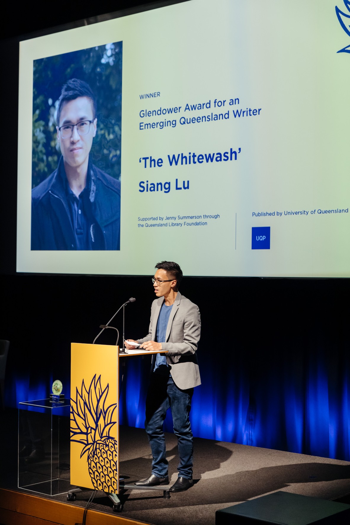 Siang Lu stands onstage behind a lectern with a blue and yellow pineapple on the front. Behind him is the name of his winning novel onscreen, 'The Whitewash'