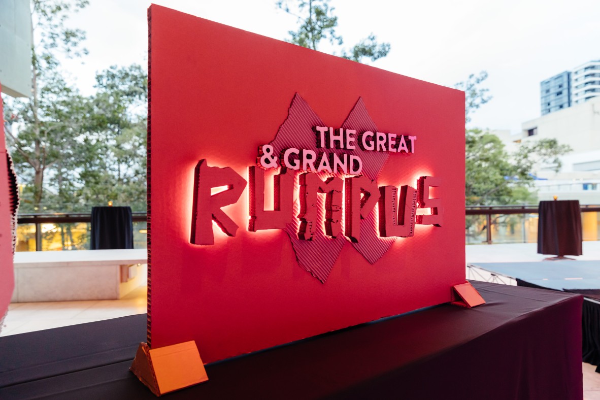 Photograph of an illuminated cardboard sign that reads The Great  Grand Rumpus The sign is red and the words are in lights   