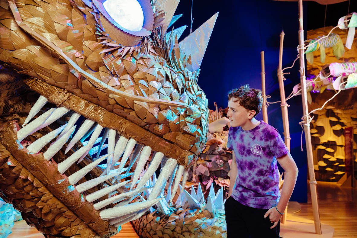 A young boy is standing close to a large cardboard dragon 