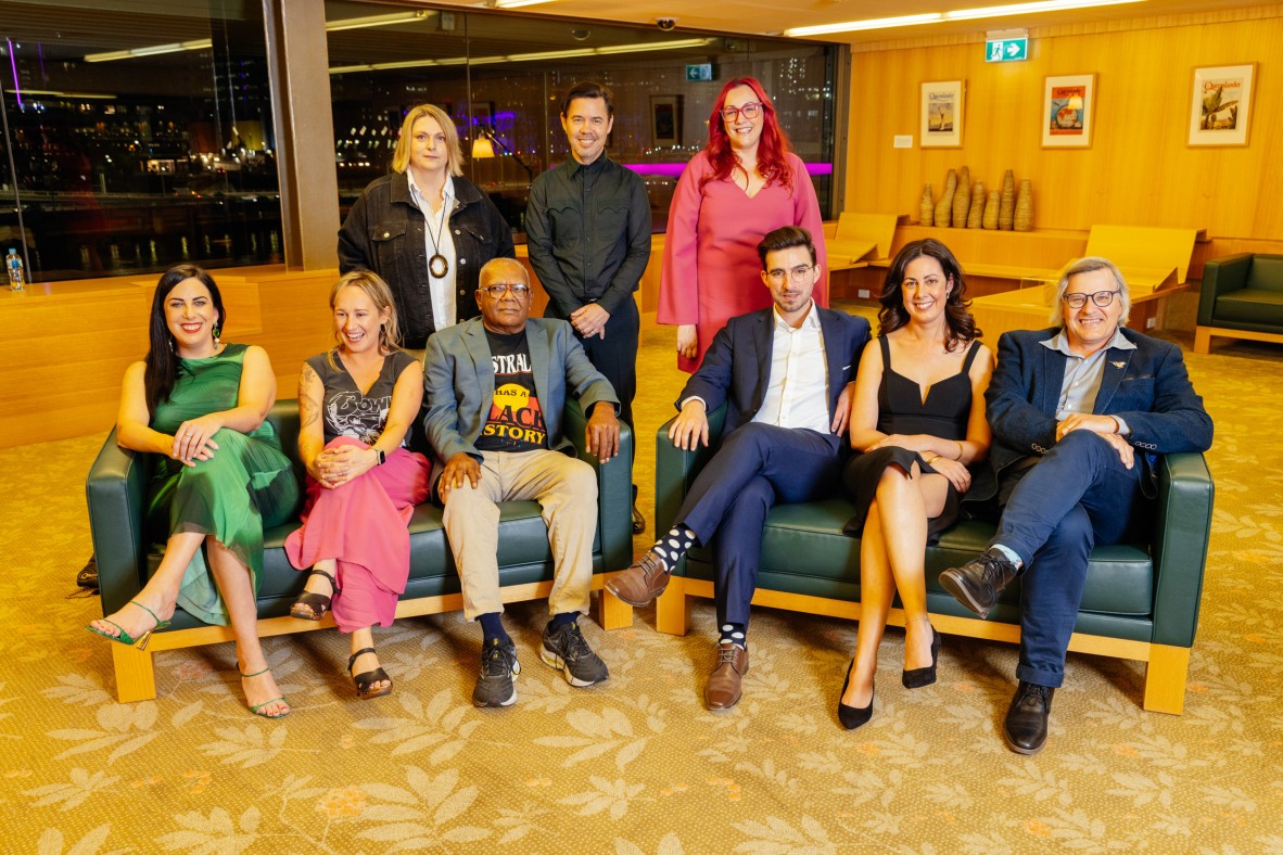9 writers stand and sit on couches in John Oxley Library at State Library of Queensland. Outside the windows is the city at night.