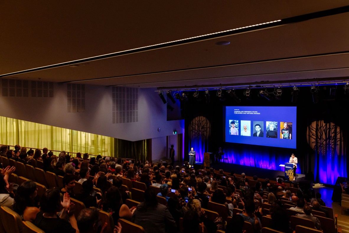 Large auditorium of people watching someone speak on stage with a projection behind them.