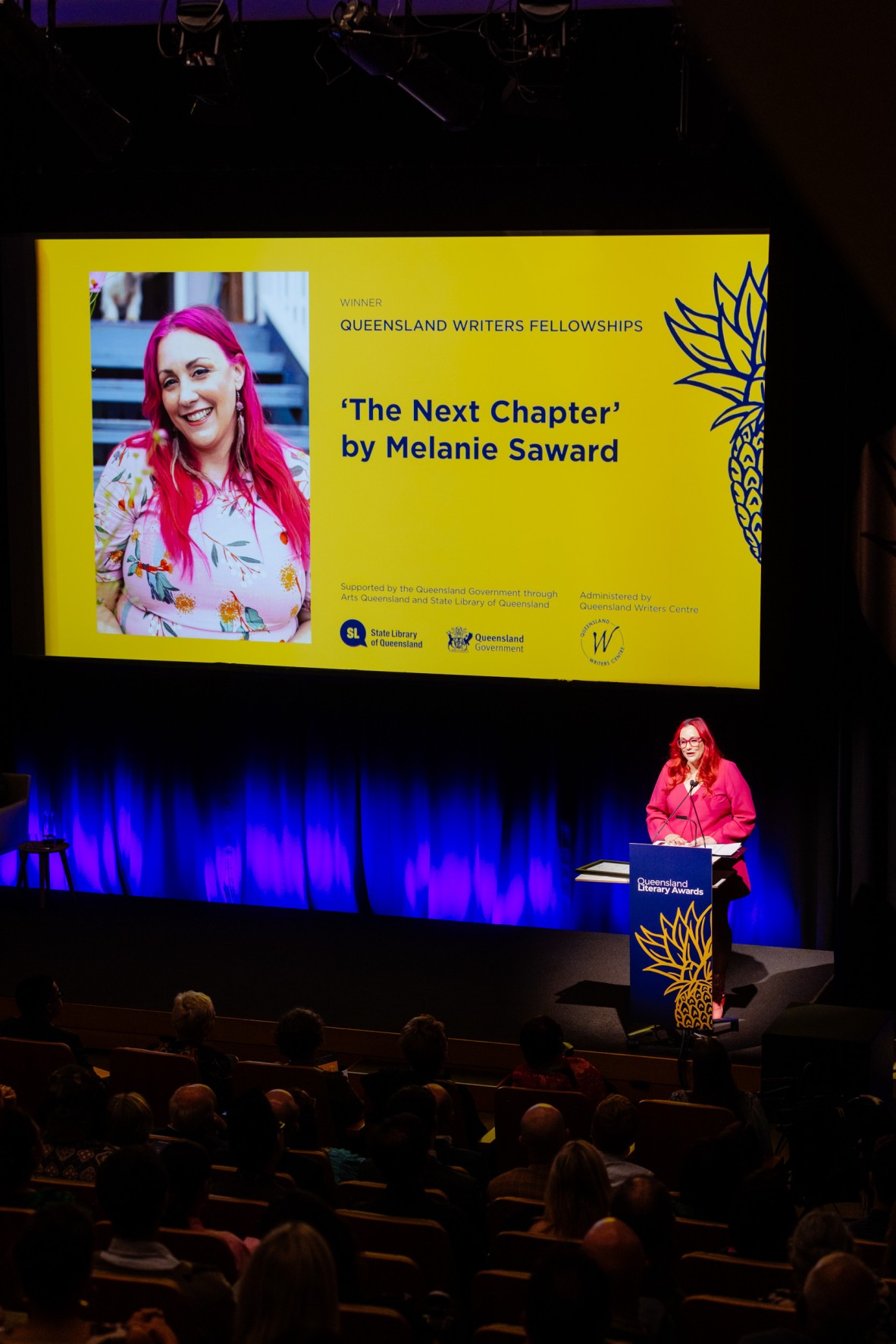 A view of a stage with a woman in pink at the lectern and a yellow slide on the screen saying 'The Next Chapter' by Melanie Saward