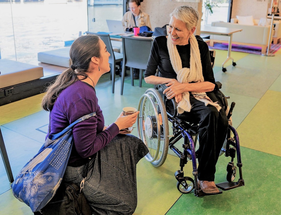 Lorraine is in a wheelchair and chats with an attendee. She is wearing a beige scarf. The woman she is speaking to is kneeling to speak with her.