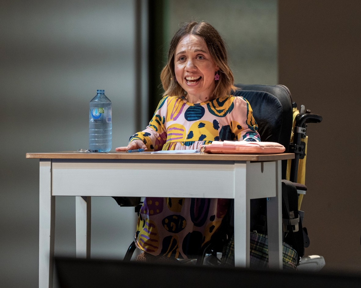 Beth wears a colourful dress and pink flower earrings. She is in a wheelchair and is talking to the audience