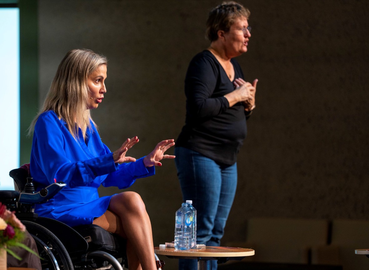 Karni Liddell wears an electric blue dress and talks to the audience. An Auslan interpreter is in the background