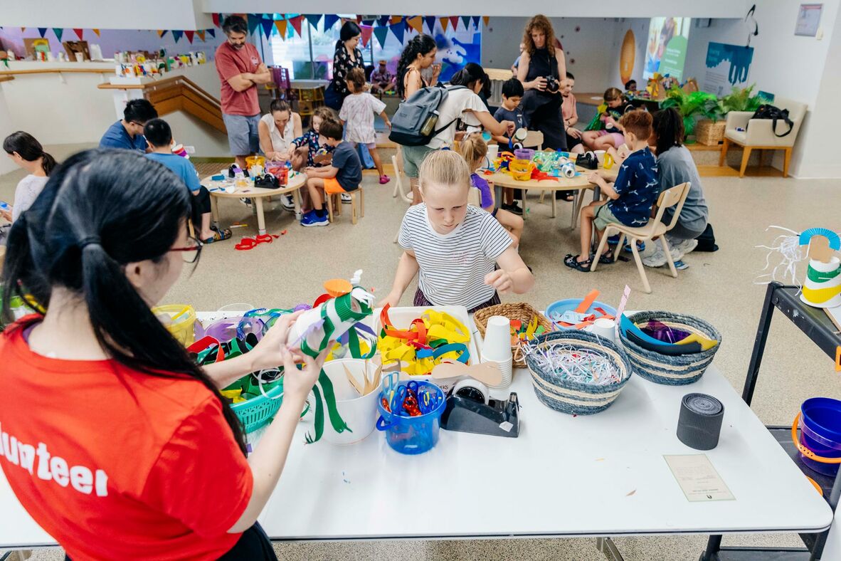 A group of people, including volunteer, children, and carers, engaged in a creative workshop at the library.