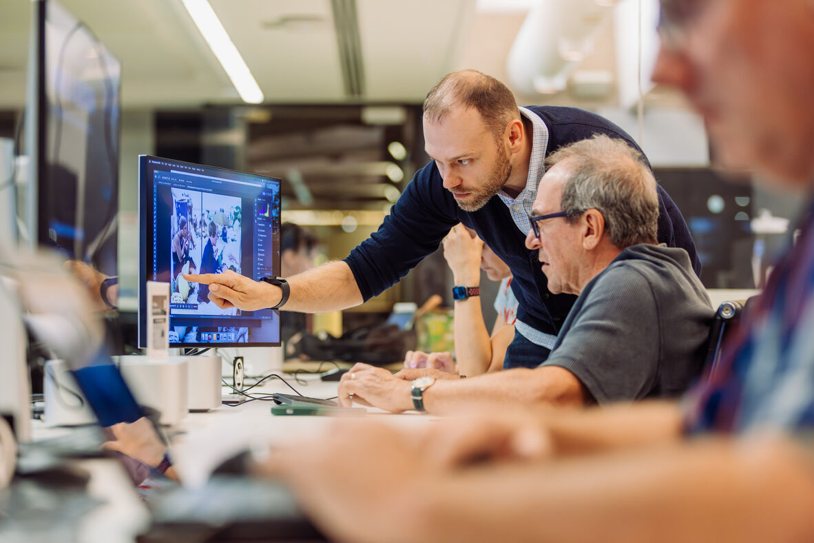 A photo of a man pointing at a computer screen showing another man what to do