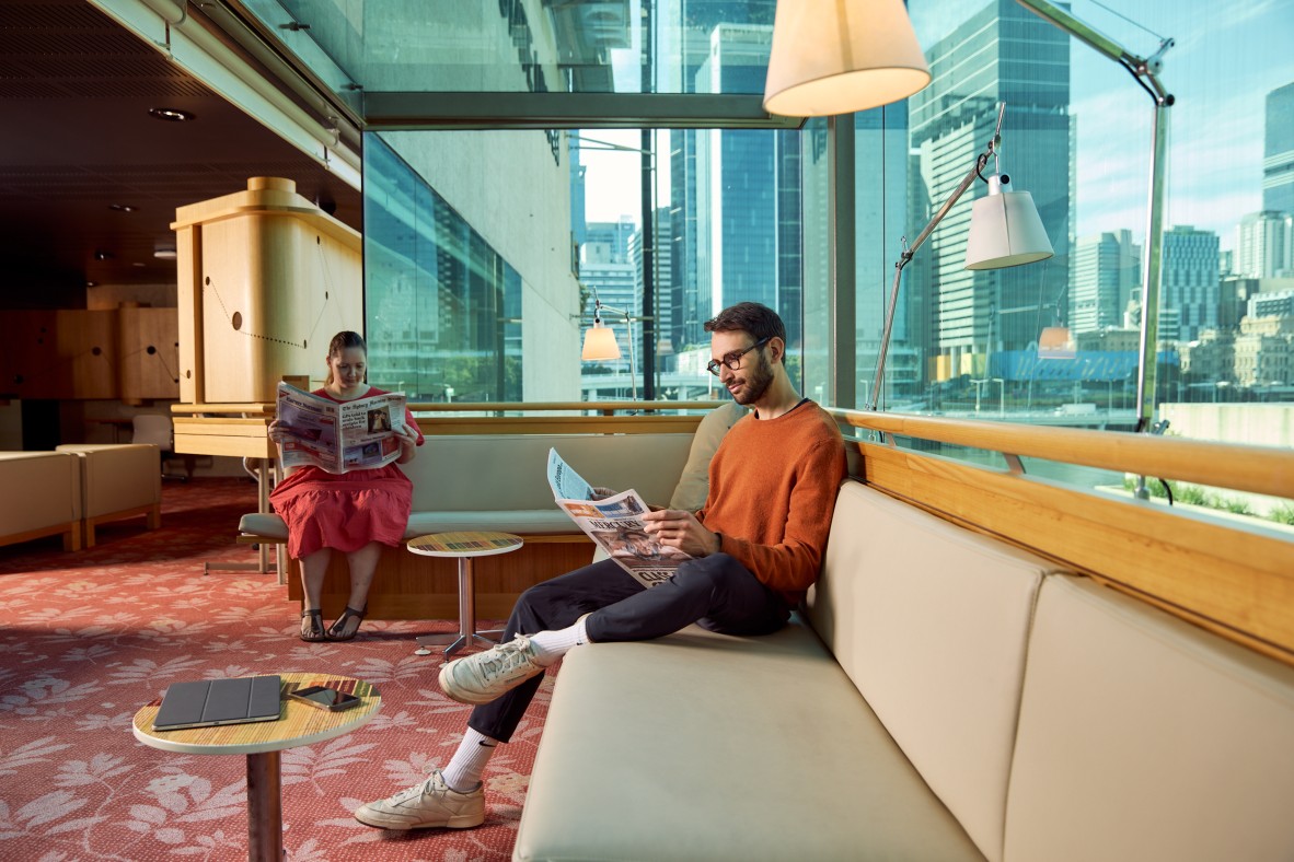 A man and woman sit in State Library of Queensland reading newspapers