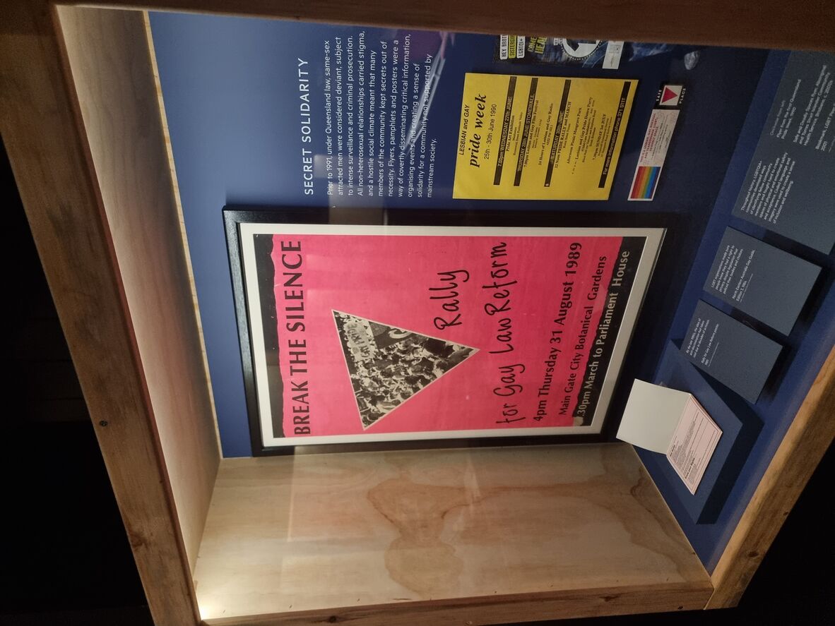 A framed poster with the title Break the Silence Rally, one Pride Week flyer, a bookmark, and a health check flyer in a lit glass display case. 