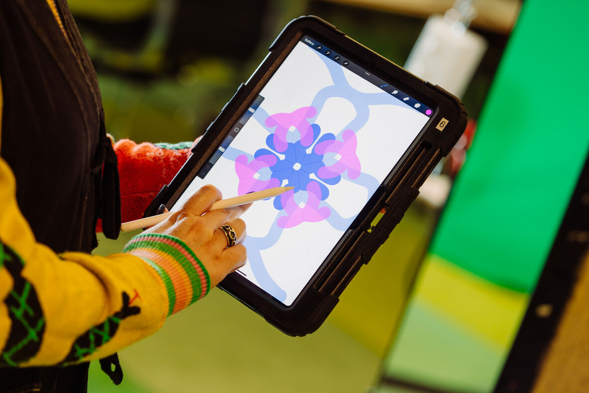 A black cased iPad can be seen being held by a person who is drawing on the screen with an Apple Pencil. Procreate is open and there are bright swirls and lines.