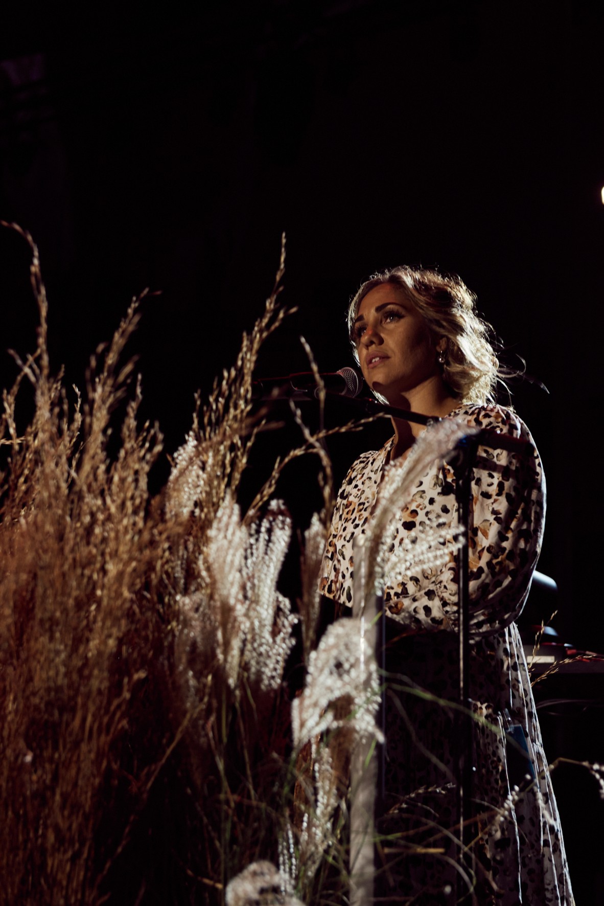 Photo of Kirli Saunders at night, standing at a microphone. Dried flowers are in the foreground. She wears a leopard print dress.