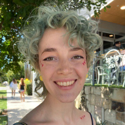 Headshot of Ellie Kaddatz She is smiling at the camera outdoors with trees behind her She has pale blue curly hair and yellow earrings