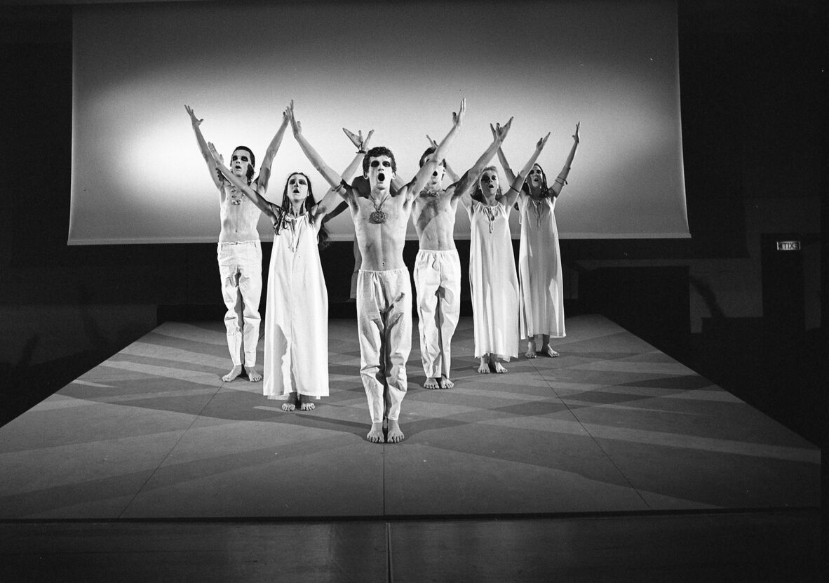 a black and white photo of 6 threate performers on stage dressed in white outfits. The men are shirtless. They are standing in an arrow formation with there hands above their head in a Y shape.