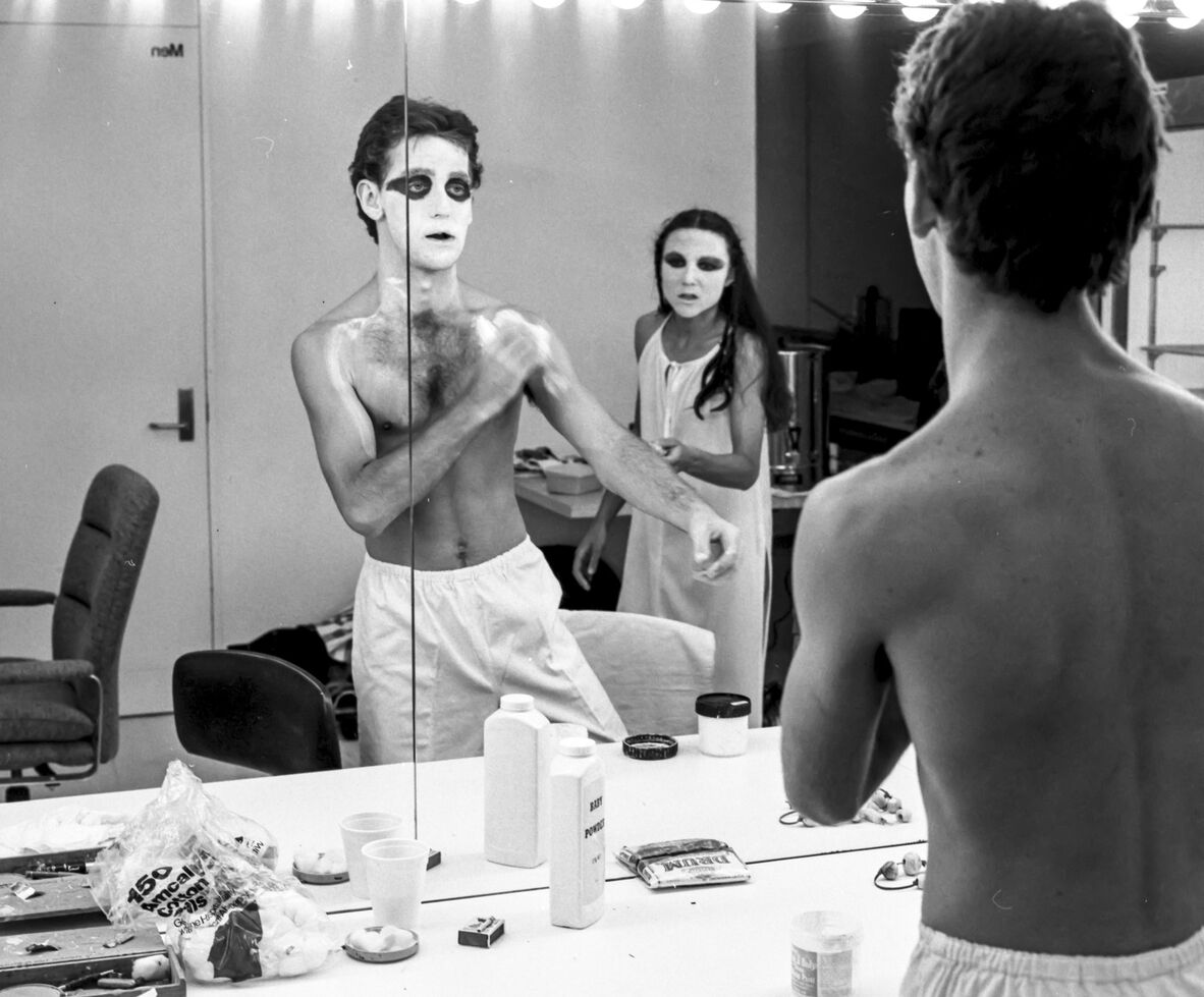 A black and white photo of a male threate performer applying white makeup in mirror. He has white facepaint on with dark. black circles around his eyes. He is shirtless and wearing white pants. A women is seen in the background of the mirror with the same facebook and a white dress.