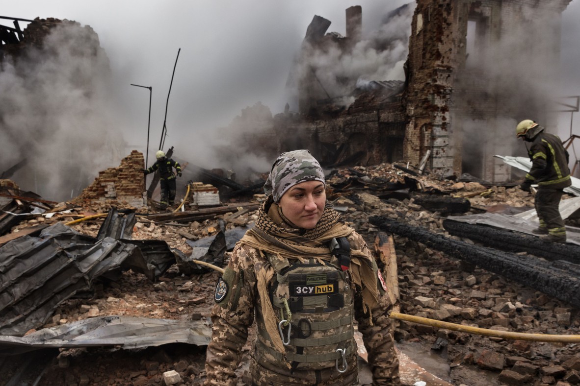 A photo of a woman in army clothing in front of bombed building in Ukraine