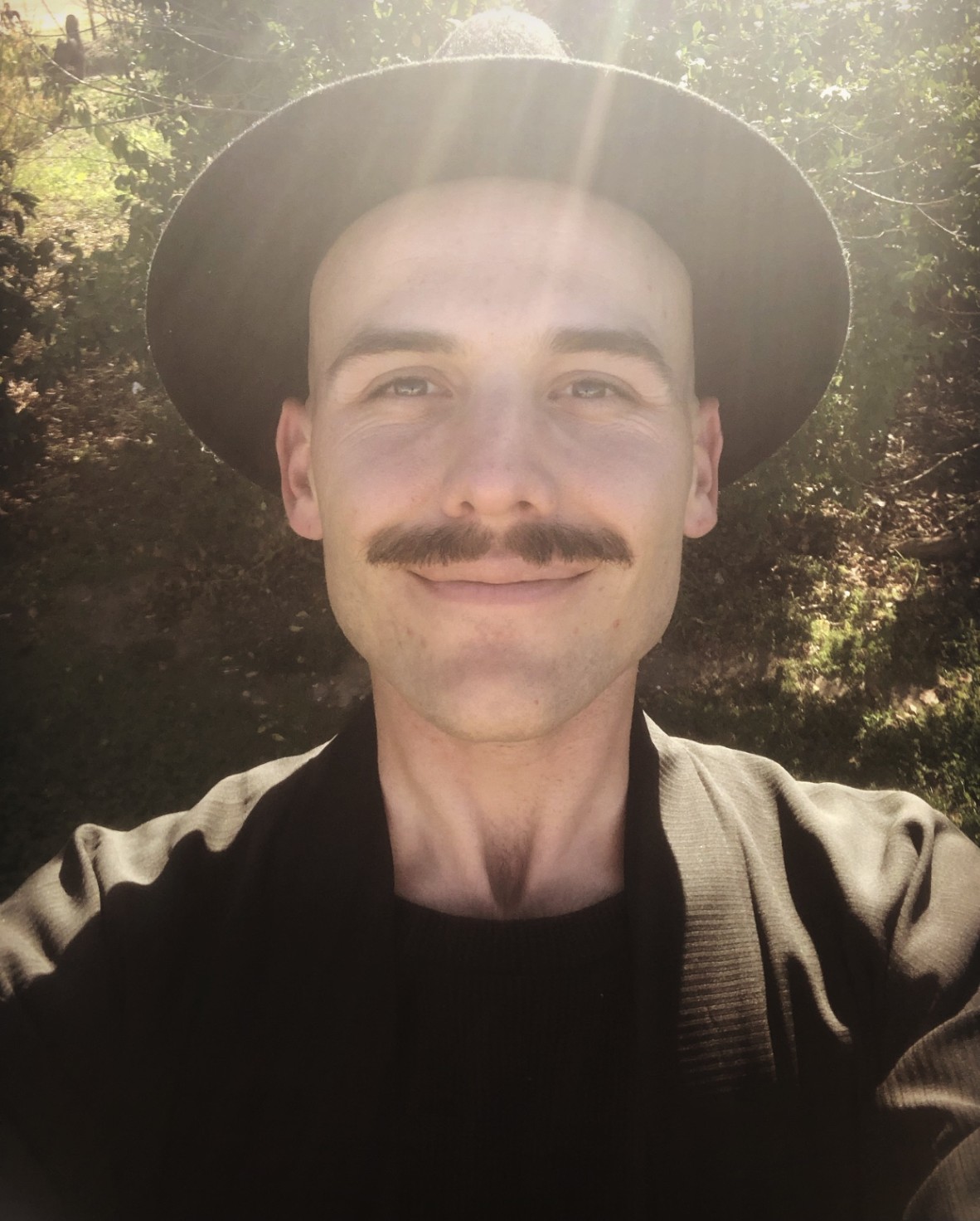 A selfie of Darby Jones who is wearing a black jacket and hat He is smiling at the camera with sun and a tree behind him