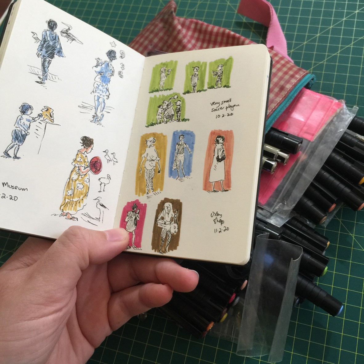 An open sketchbook held in front of a collection of illustrators pens