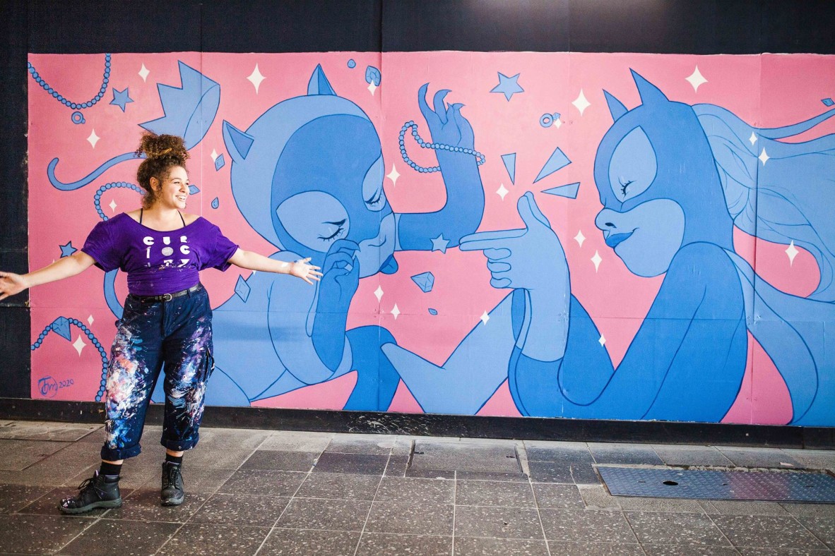 Tori-Jay Mordey stands smiling in front of a large mural “Cat Gal vs Bat Gal” – it is pink and blue