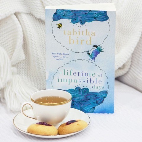 A copy of A Lifetime of Impossible Days sits against a white blanket, beside a cup of tea and biscuits