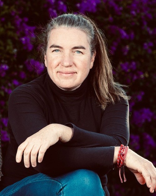 Photo of A E Macleod They are seated with arms crossed over wearing a black top and in front of a purple flowered background