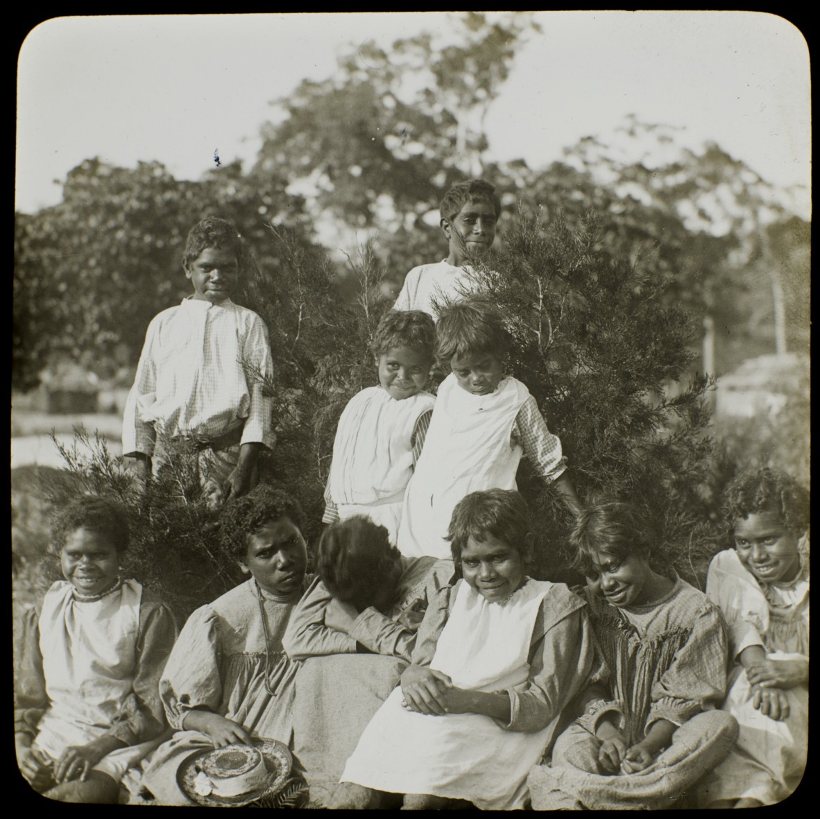 Group of First Nations children in front of shrubs, four children are standing with six children sitting on the ground in front.