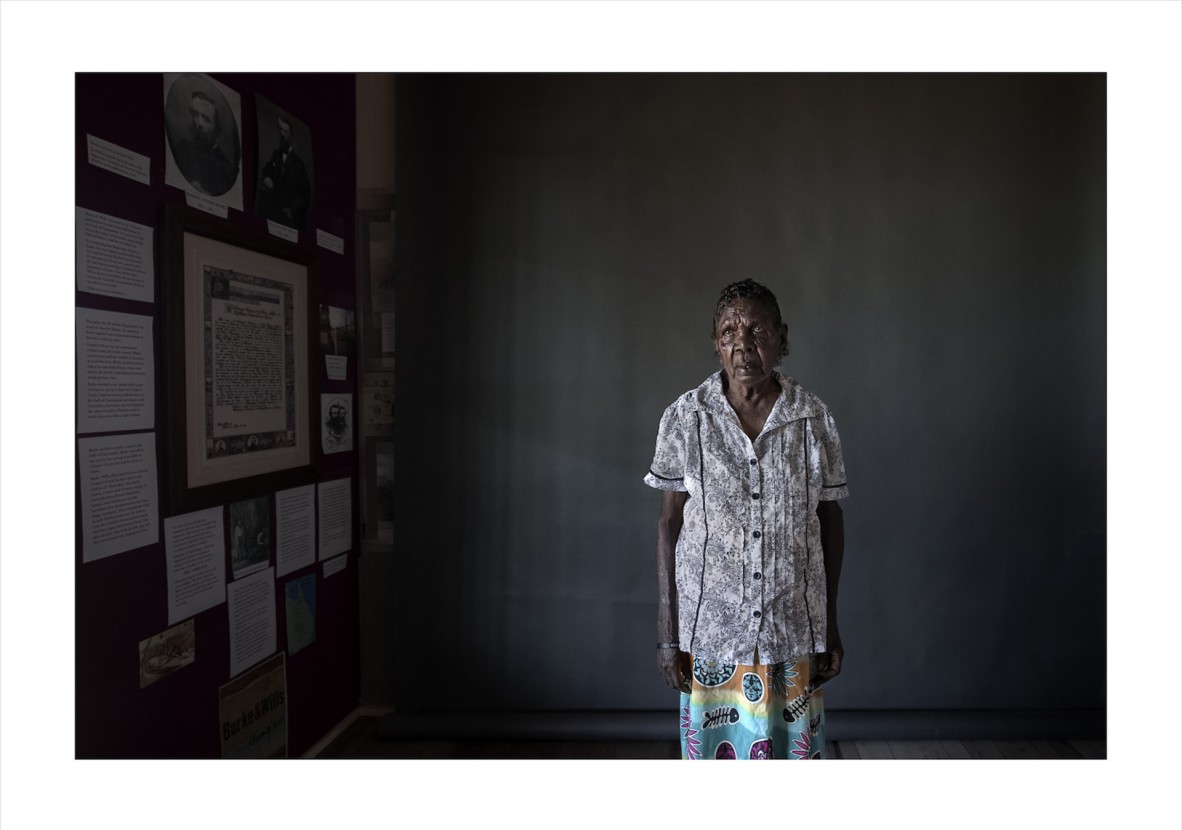 First Nations Elder standing near portraits on a dark grey wall.