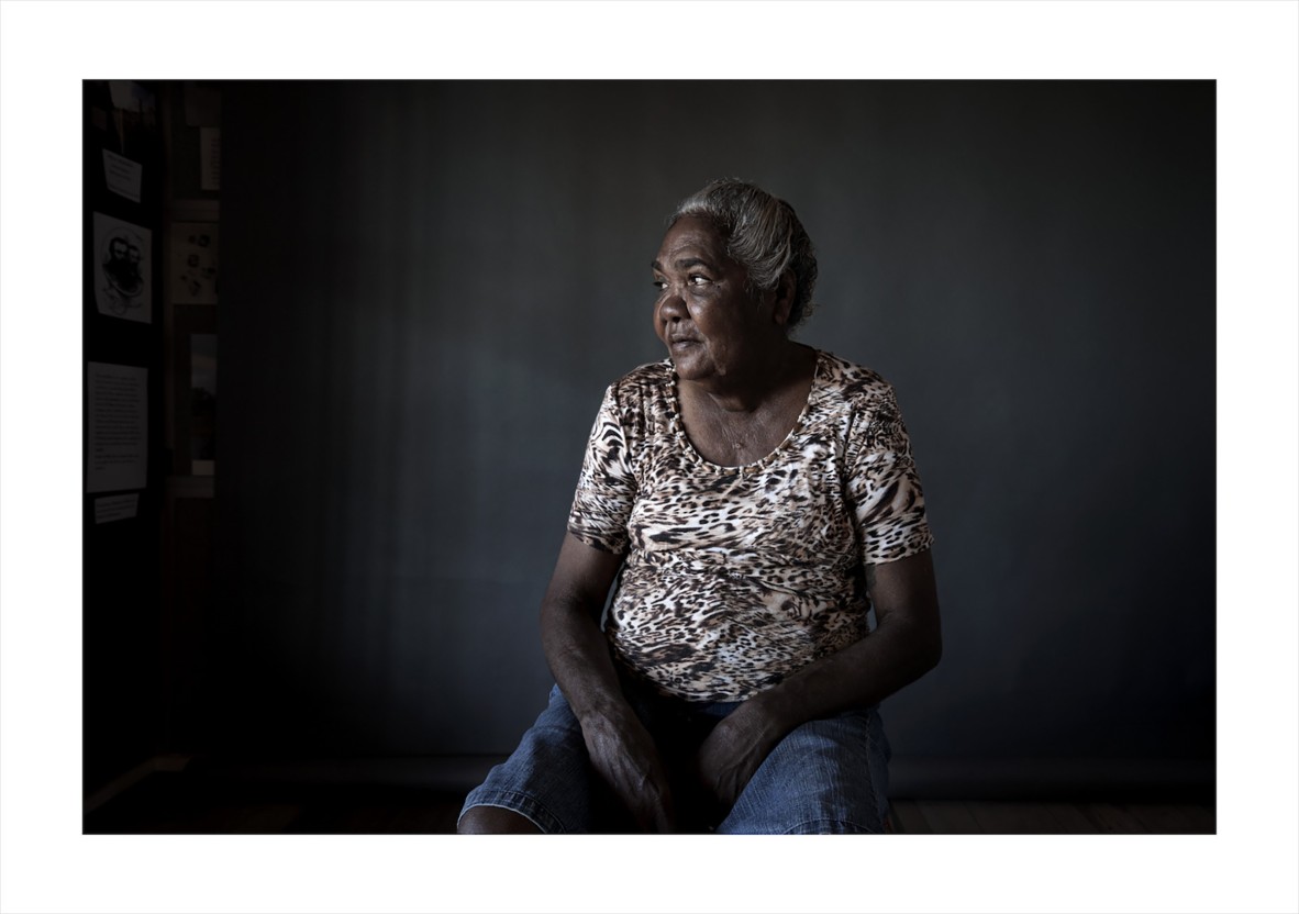 First Nations Elder sitting on stool against dark grey wall. 
