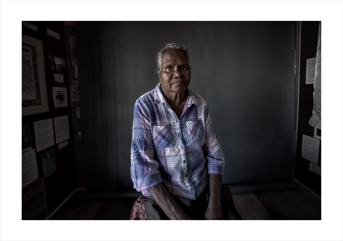 First Nations Elder sitting on stool against dark grey wall.