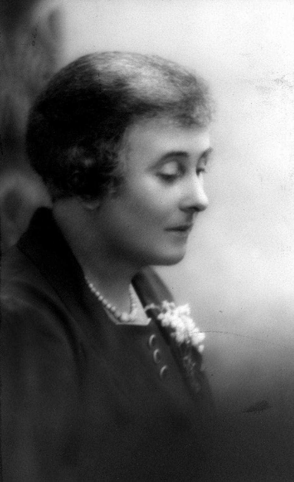 A side profile of a woman wearing pearls and a flower pinned to her jacket