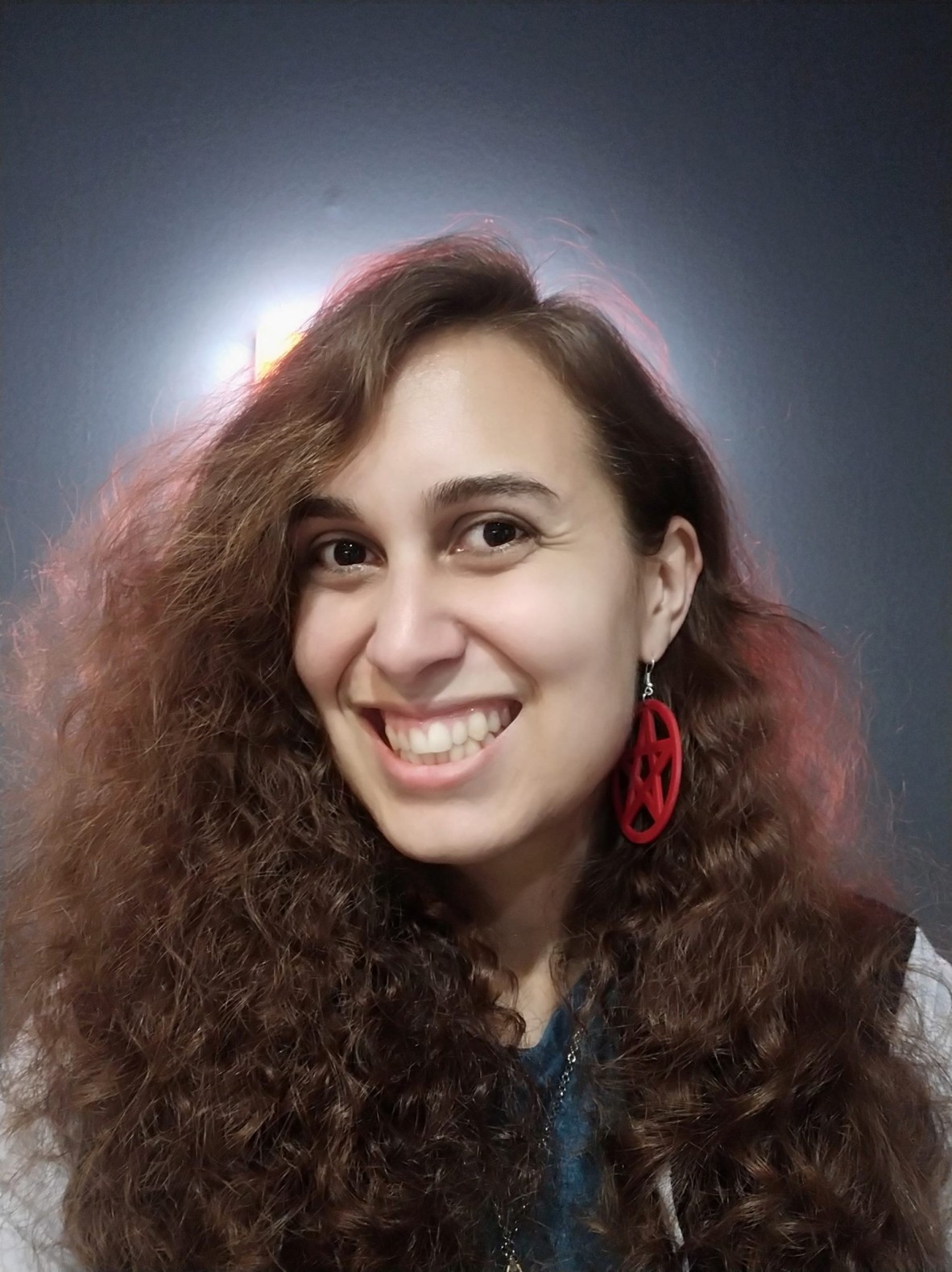 A photograph of writer Allanah Hunt who has wavy brown hair haloed by soft lighting and wears red pentagram earrings