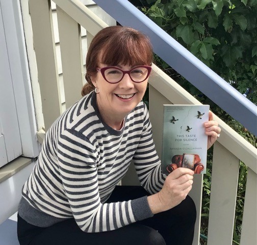 Amanda O'Callaghan sits on the steps outside her house. She wears a grey and white striped shirt, dark red glasses, and holds up a copy of This Taste for Silence.