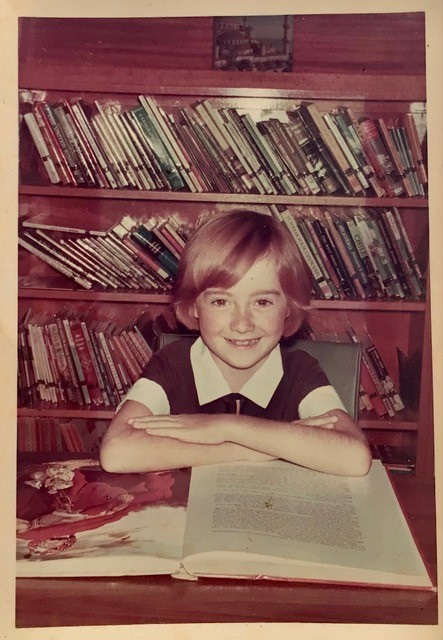 A small girl with short hair sits and smiles in her school uniform with shelves of library books behind her. She is at a desk with her elbows on an open book.