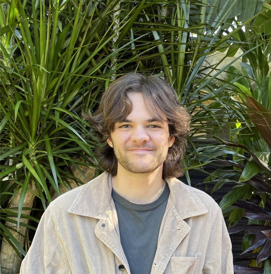 Photo of Ash Shirvington who stands smiling in a garden. He is wearing a brown shirt.
