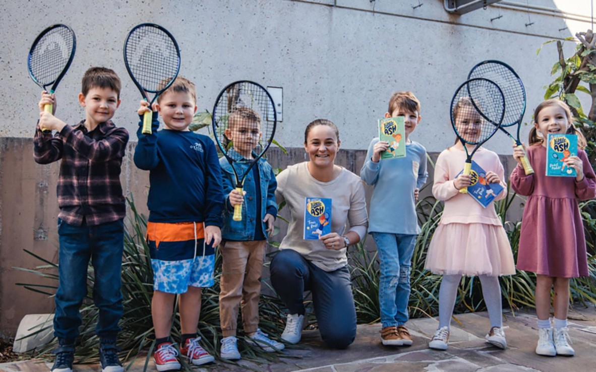 Ash Barty, Australian tennis player, kneeling with a group of children holding tennis rackets and books.