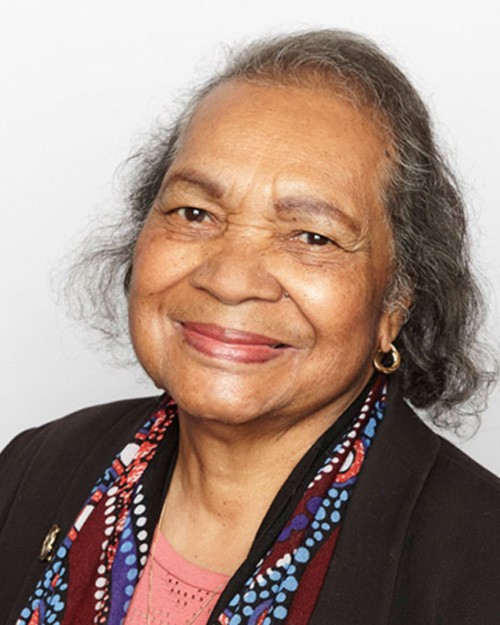 Headshot of Aunty Joan Tranter. She is standing against a white wall and is wearing a black blazer, colourful scarf and pink shirt