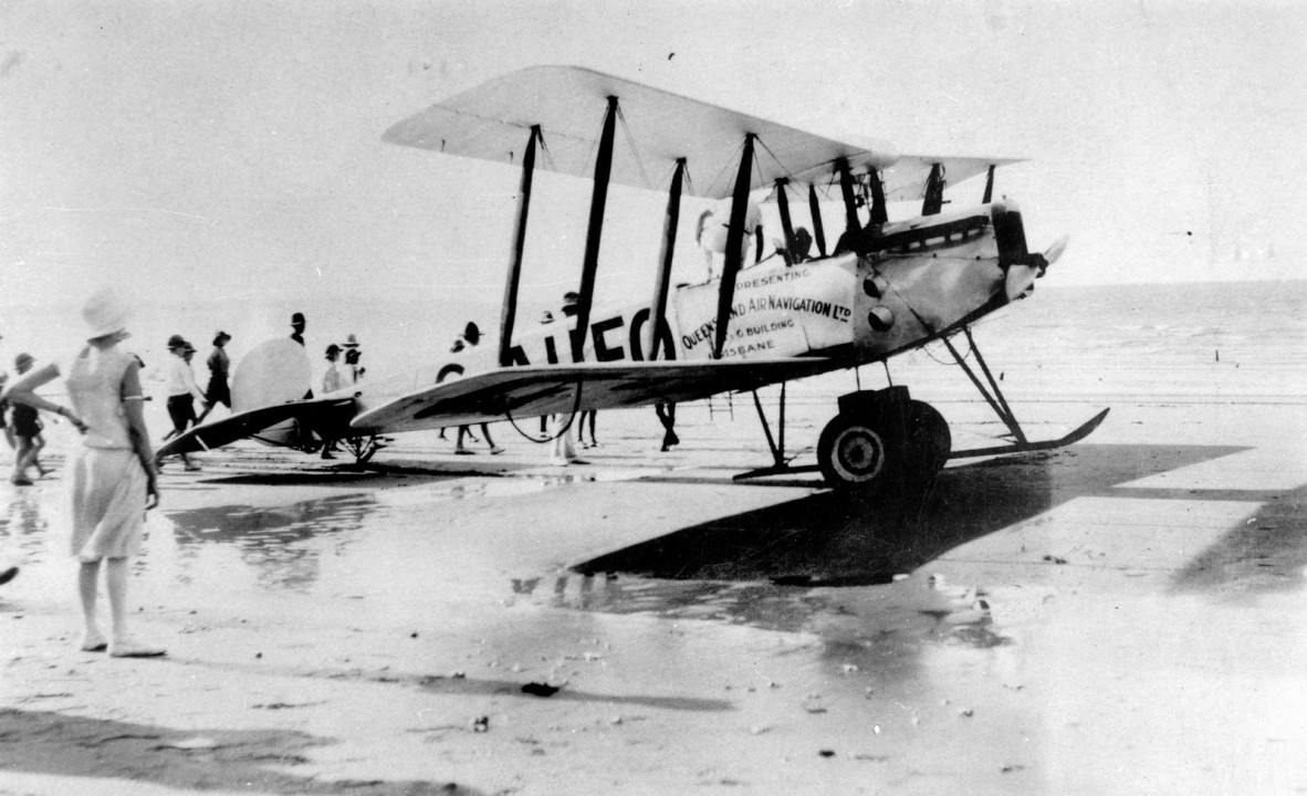 Avro 504K biplane G-AUEO with an advertising sign on the side saying Queensland Air Navigation Ltd., Brisbane. The plane has landed on the beach and a crowd of onlookers have gathered to inspect it.