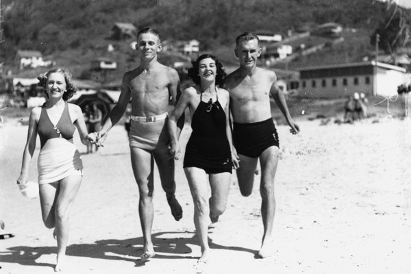 A party of beachgoers runs across the sand. They wear a variety of swimwear.