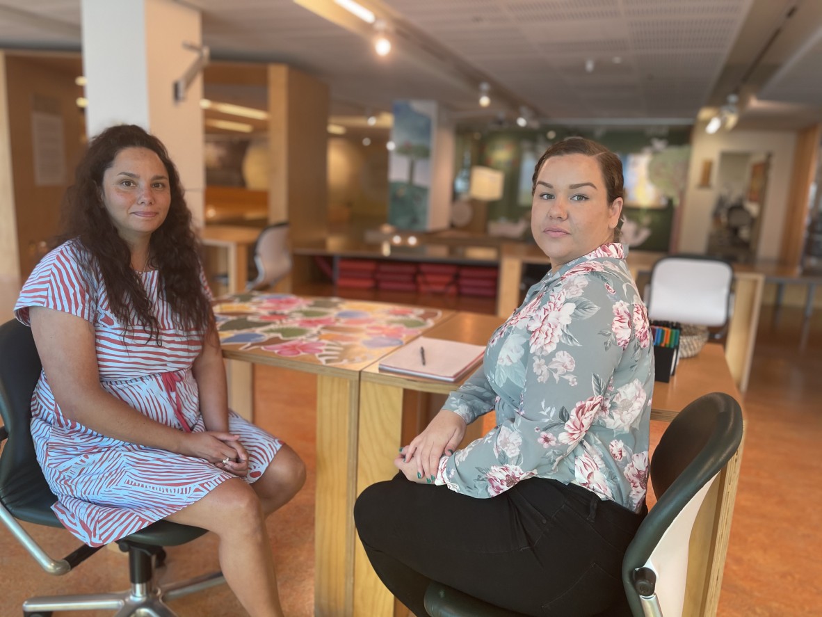 Bianca Valentino and Nadia Johansen sit at a desk in kuril dhagun, facing the camera, a notebook and pen on the table between them