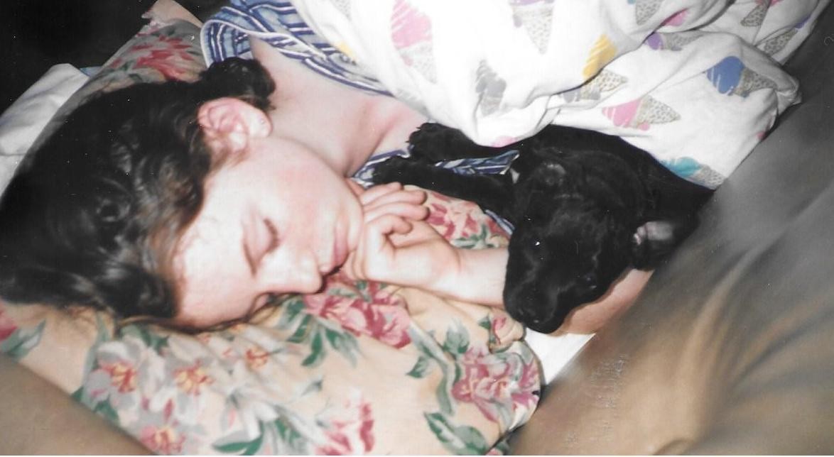 A woman is asleep on a floral cushion with a sleeping black puppy beside her