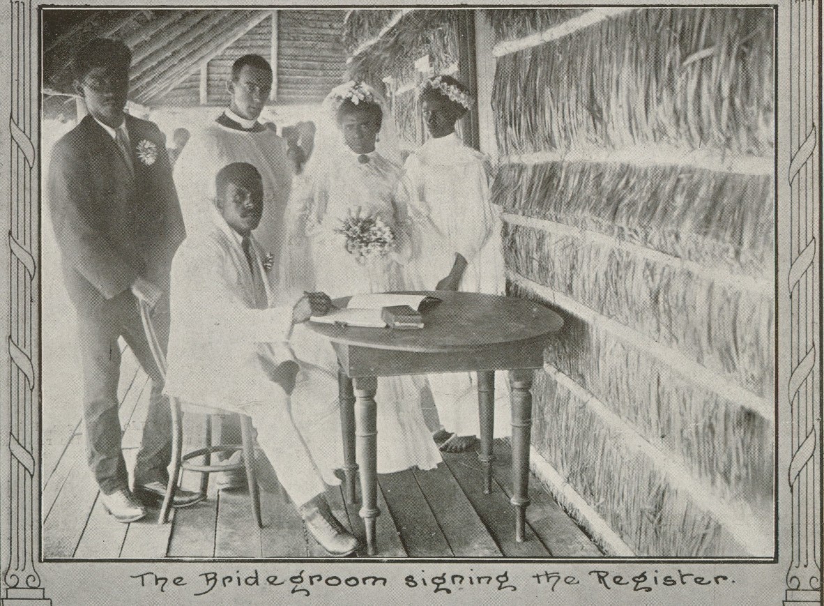 First Nations man in white suit sitting at a table with four First Nations people standing behind him: two men and two women in white dresses