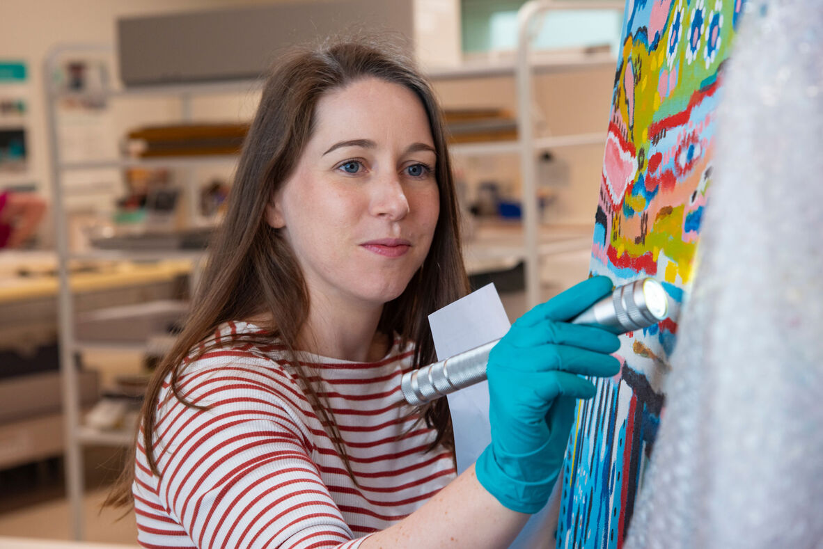 Woman with long brown hair and striped shirt shining torch on colourful First Nations painting