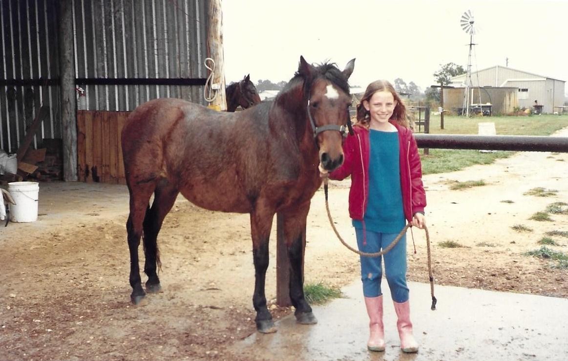 A smiling girl stands next to a brown horse on a farm