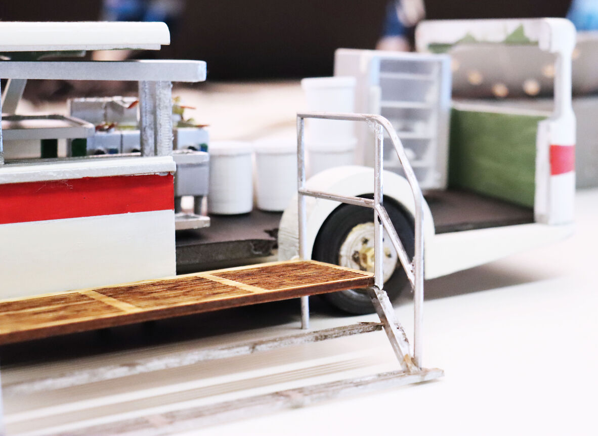 A close-up of a miniature model of a food truck with a vintage-inspired design. The model features a small serving counter with a red and white color scheme, an open side door, and interior details like shelves and containers. A narrow set of metal steps with a wooden platform leads up to the serving area, adding to the truck’s realistic, retro aesthetic.
