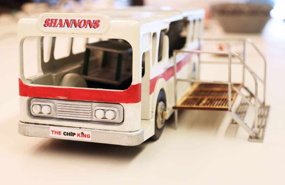 a photo of a front on model of a bus/van that is white with a red swipe. There is no visible glass and you can clearly see into the bus. There is a sign that states "Shannons". There is also some metal stairs and a platform on the right hand side.